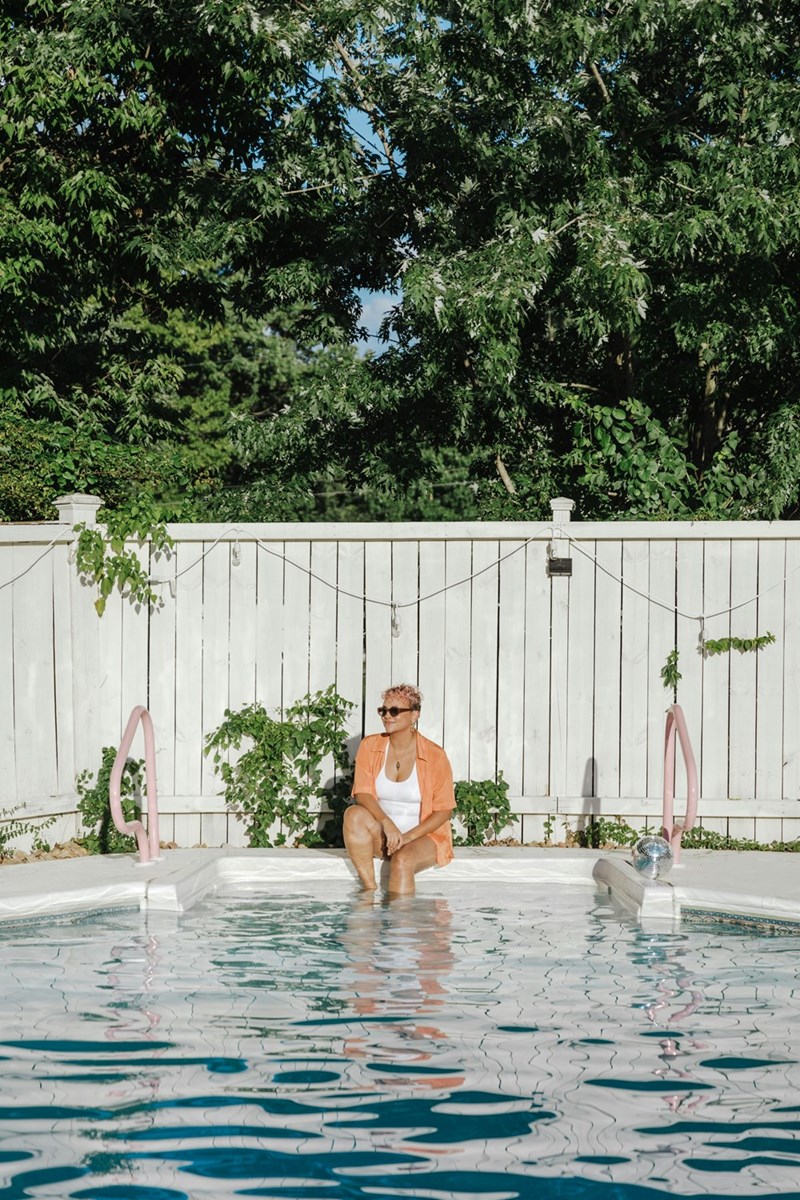 Woman in sunglasses sitting at the edge of a backyard pool with her feet in the water, against a white fence.