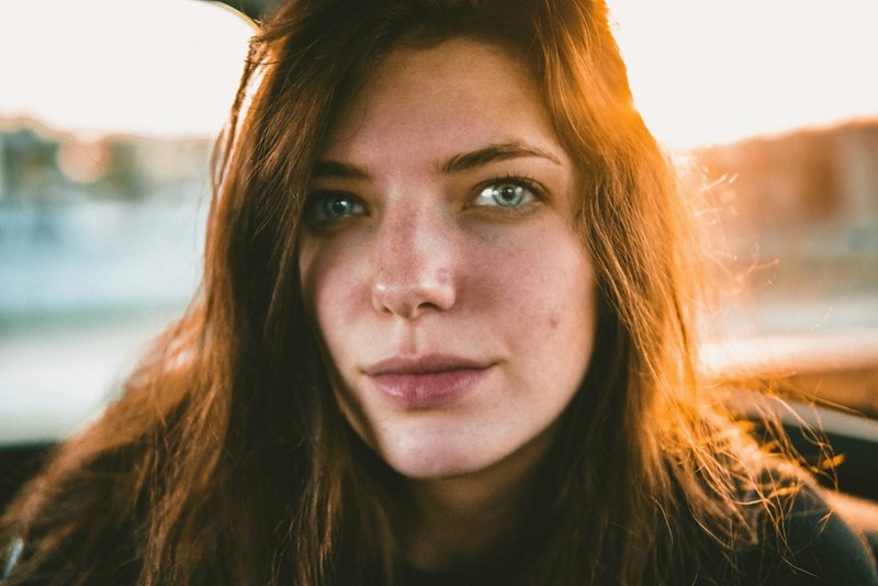 Close-up portrait of a woman with long hair and freckles, lit by warm sunlight in the background.