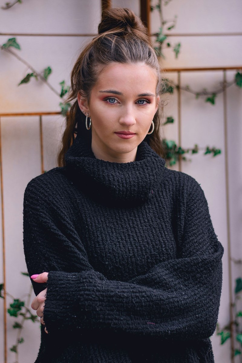 Portrait of a young woman with her hair in a high bun wearing a black turtleneck sweater, standing with arms crossed against a light wall with greenery.