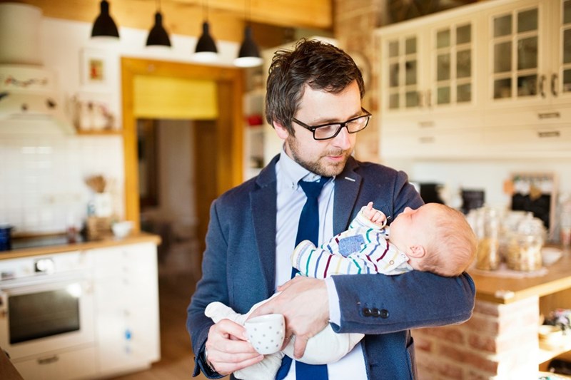 Young businessman coming home from work holding his sleeping little baby daughter in his arms drinking coffee