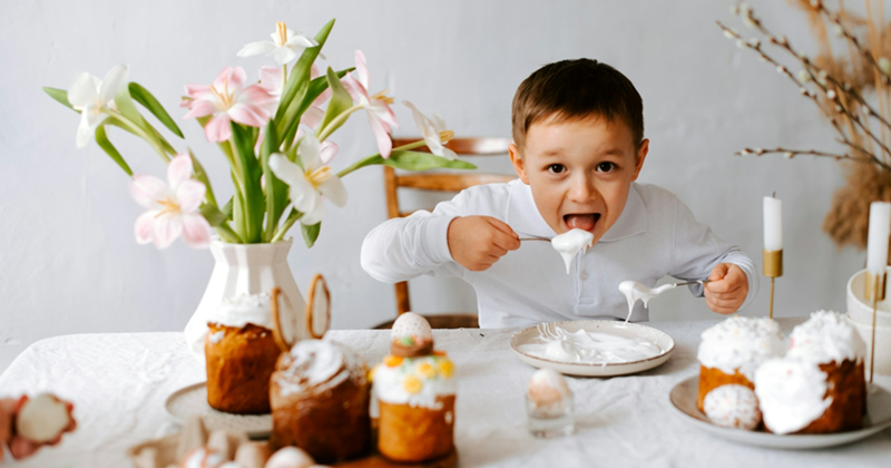 A little boy sitting at a table decorated with plates of a cakes and a bouquet of flowers eats icing from a plate