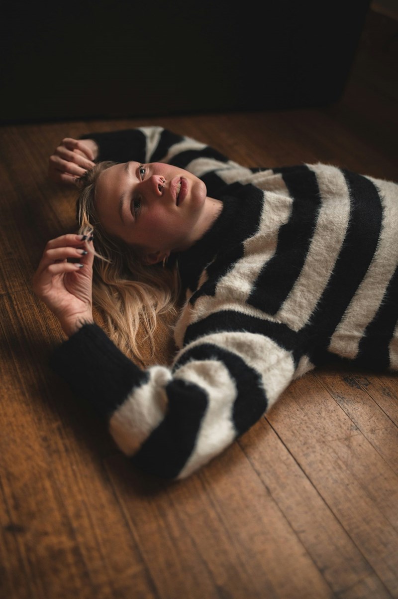 Young woman in a striped sweater lying on a wooden floor, looking upward with a relaxed expression.