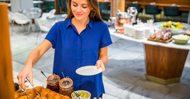A woman holding a plate uses tongs to pick up a pastry from a breakfast buffet