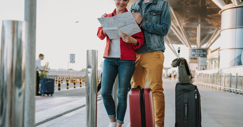A couple stands outside an airport with their suitcases, looking at a map