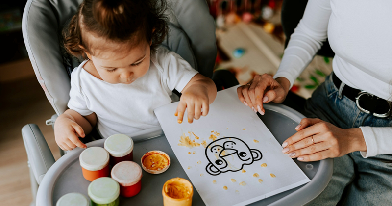 A little girl sitting in a high chair uses finger paints on a piece of paper as her mom sits beside her