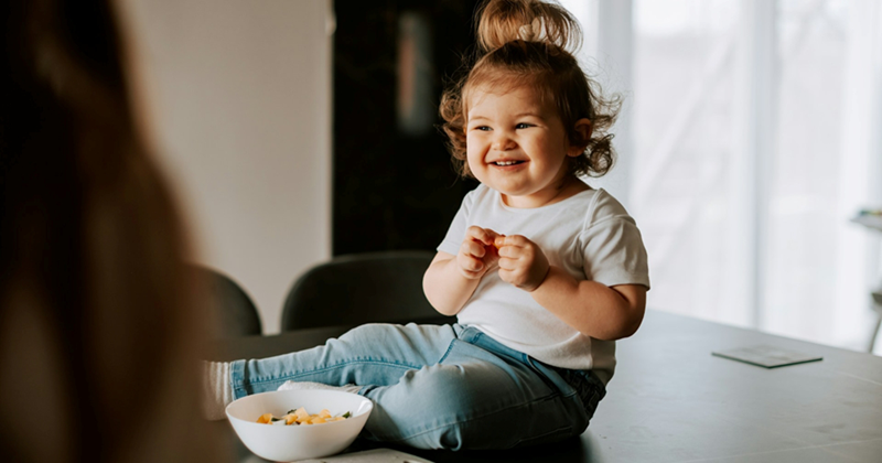 A little girl smiles as she sits on a table and eats cereal