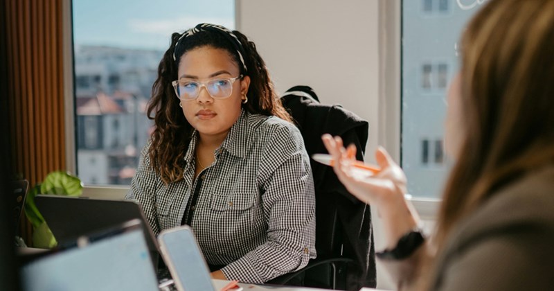 A female employee sitting at a meeting table with a tablet looks towards a female coworker who is talking