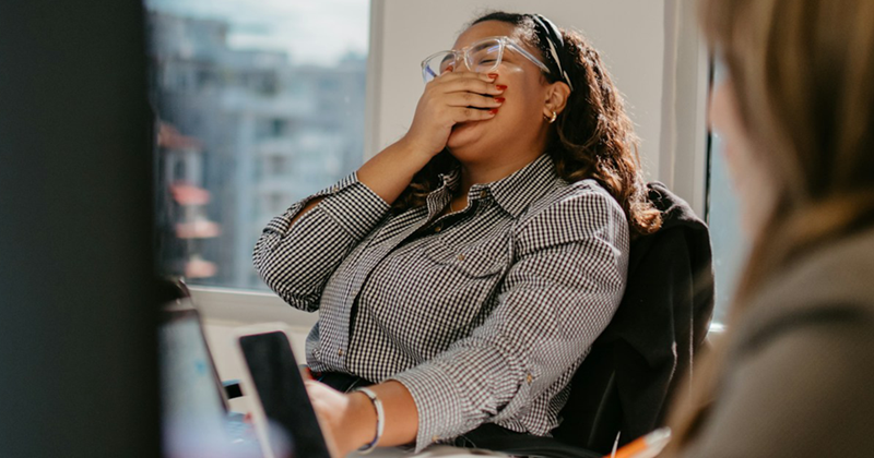 A female employee sitting at a meeting table with a female coworker covers her mouth as she laughs