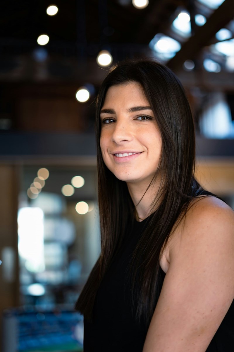 Smiling woman with long dark hair wearing a black sleeveless top, standing indoors with soft background lights.
