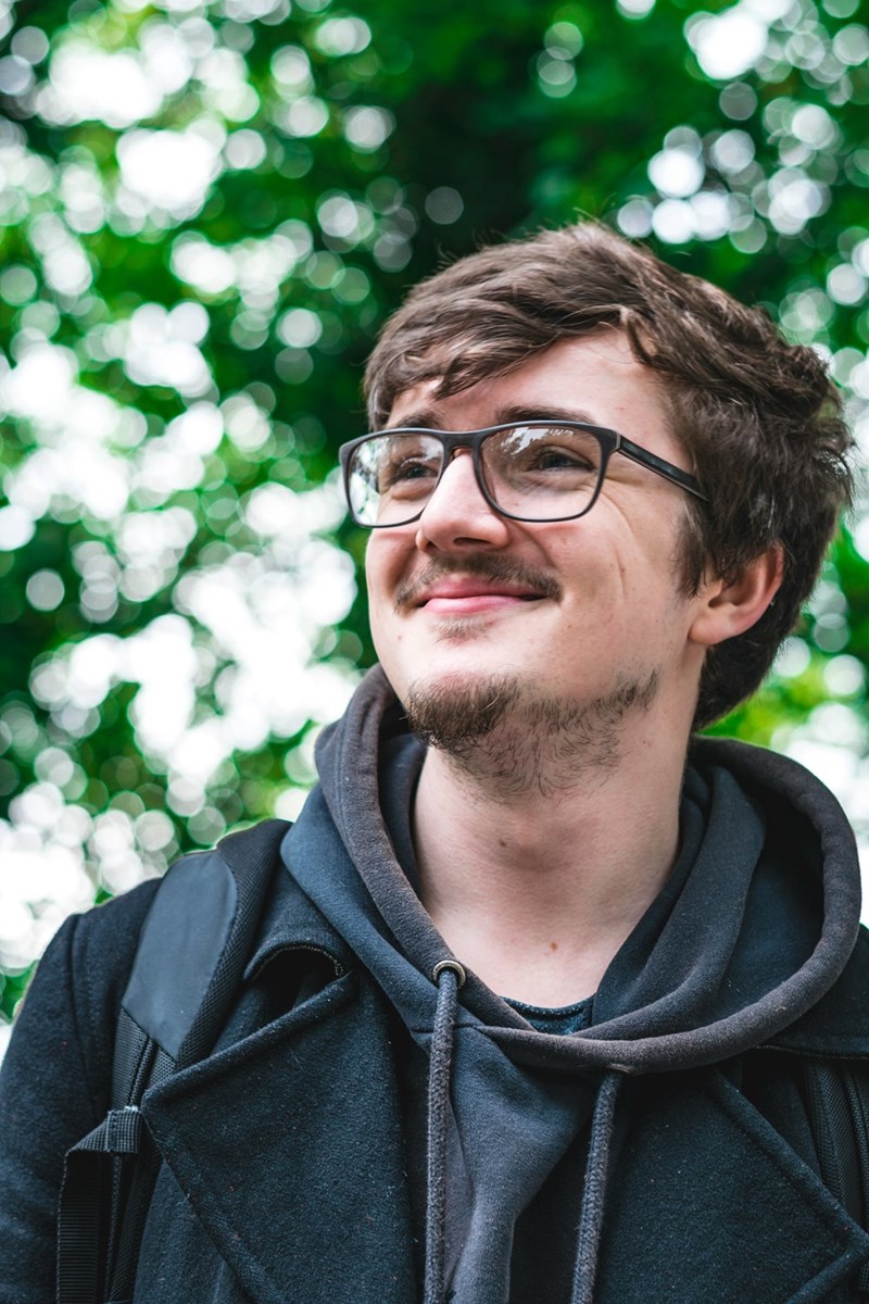Smiling man with glasses wearing a hoodie and coat, standing outdoors with green foliage in the background.