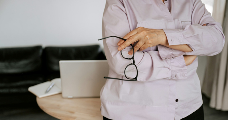 A businesswoman crosses her arms while holding her glasses, standing in front of a couch and a table with a laptop on it