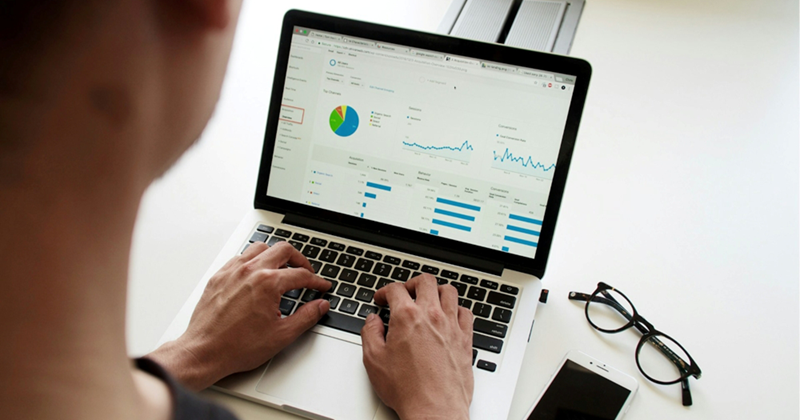 A person uses a laptop with charts and data on the screen, a phone and pair of glasses sitting next to it