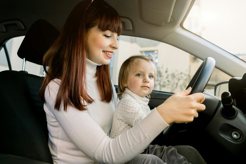 Smiling mom and her baby sit in the driver's seat of a car.
