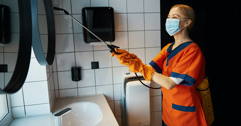 A woman in a cleaning uniform disinfects a mirror in a public bathroom with a spray canister machine
