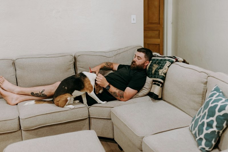 Man lying on a couch with a dog sitting on him while he holds a pen in a living room.