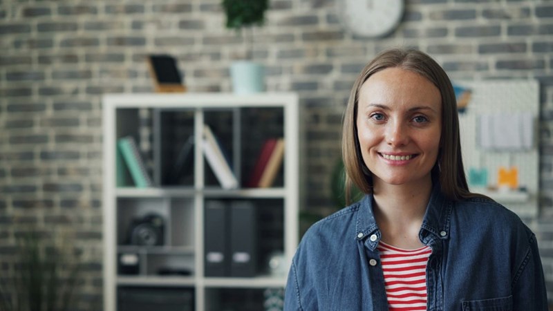 Smiling woman standing in a home office with a brick wall and shelves in the background.