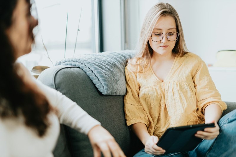 Woman wearing glasses sits on a couch holding a tablet while another person sits nearby, suggesting a conversation or session.