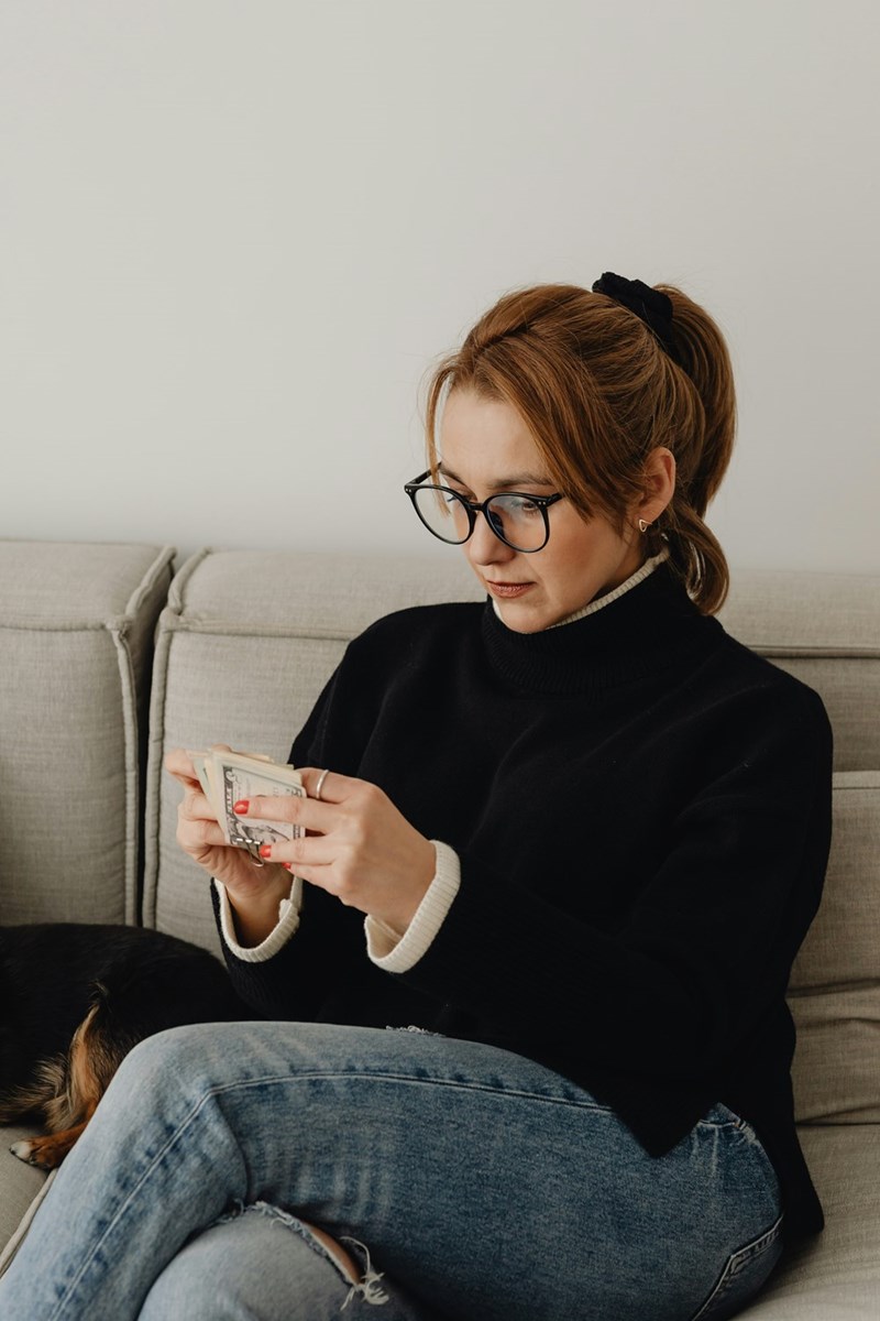 Woman wearing glasses sits on a couch counting cash, with a dog resting beside her.