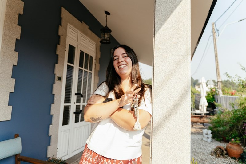 Smiling woman standing on a porch holding a set of keys, leaning against a column outside a house.
