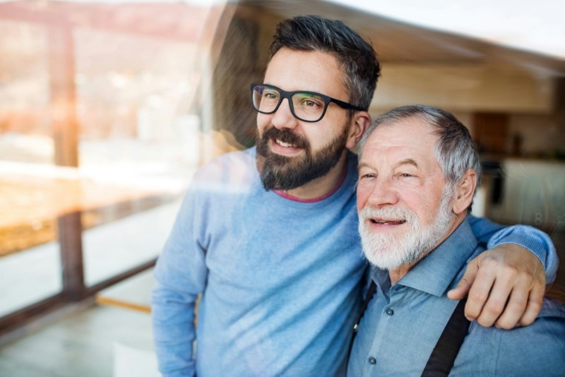 An adult man embraces his retired father as they look out the window together.