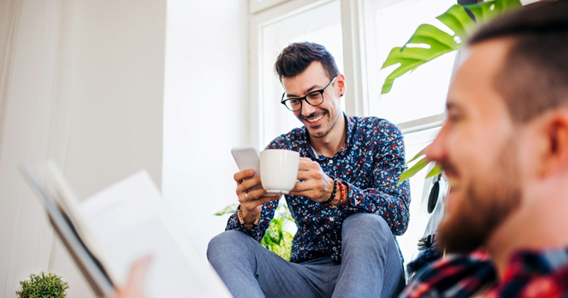 A man sits on a windowsill holding a mug and checking his phone, while a bad blurred in the foreground sits and reads