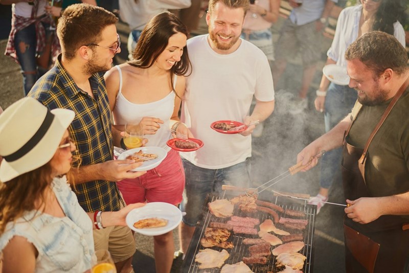 Group of people standing around grill, chatting, drinking, and eating
