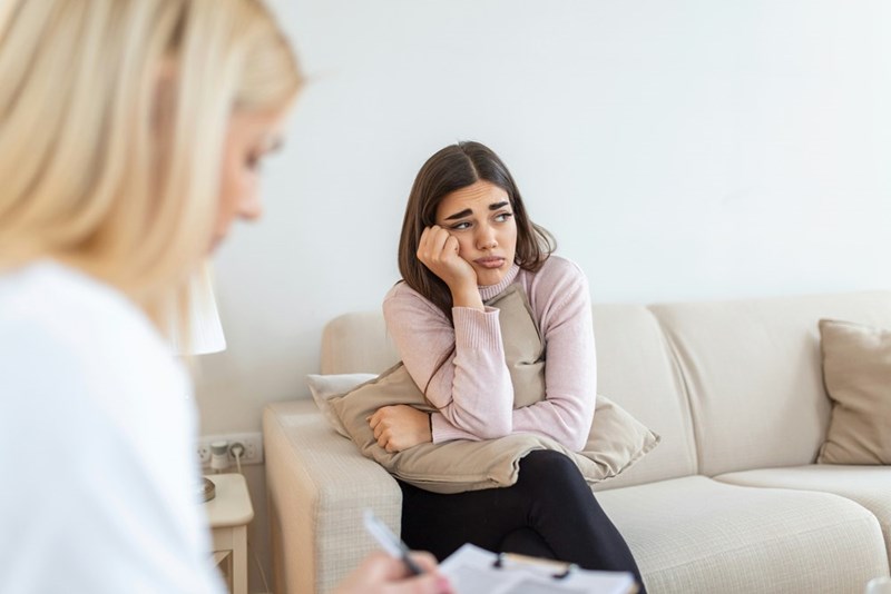 A woman sits on a couch looking upset while speaking to someone, resting her head on her hand.