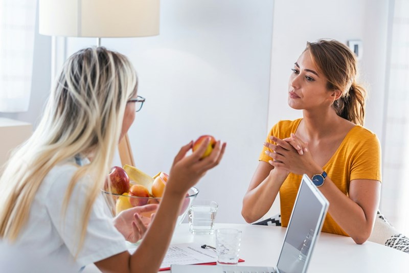 Two women sit across from each other having a serious conversation, one listening intently while the other gestures with a piece of fruit.