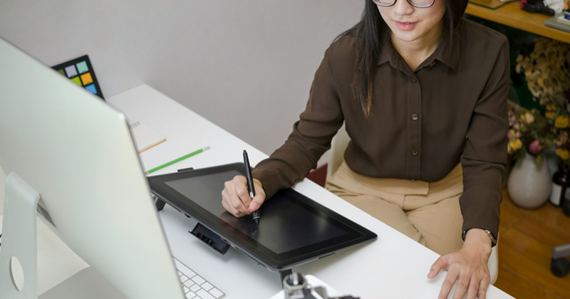 A woman in business attire draws on a tablet at a desk in front of a computer