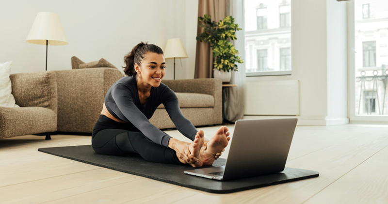 A woman wearing activewear smiles as she looks at her laptop screen and stretches on a yoga mat in a living room