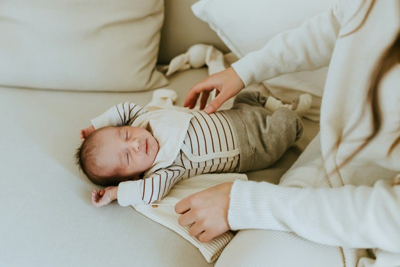 Parent sits next to infant baby, who lays peacefully on couch.
