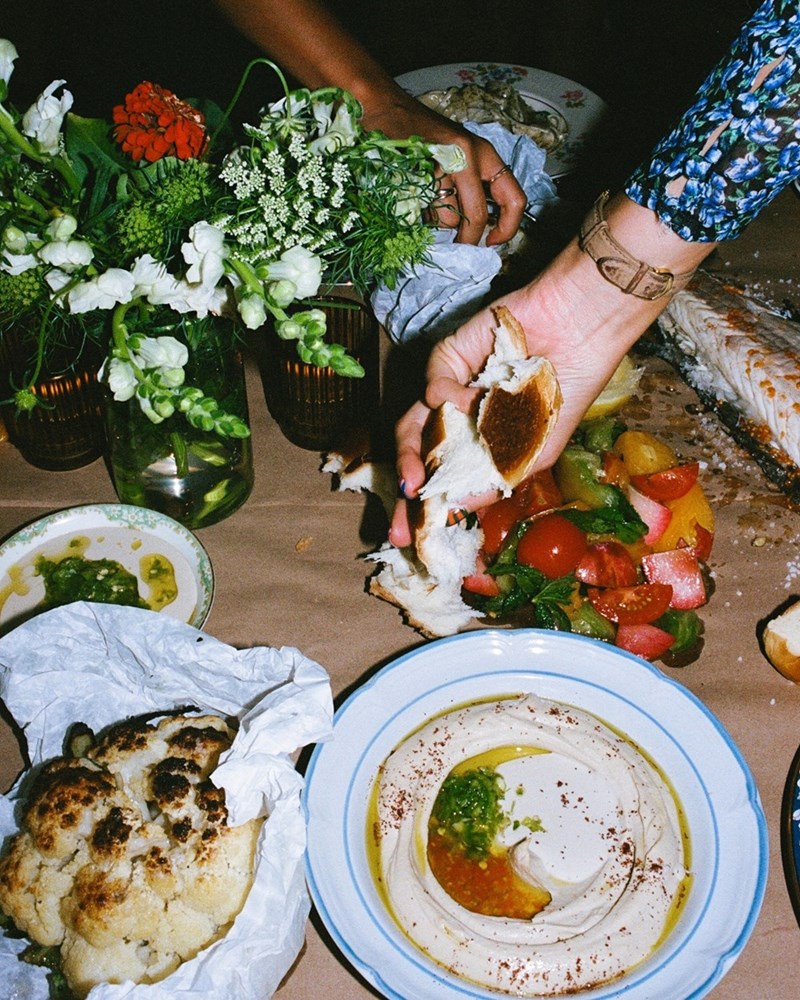 A woman's hand grabs a piece of torn bread above a table with flowers and food on it