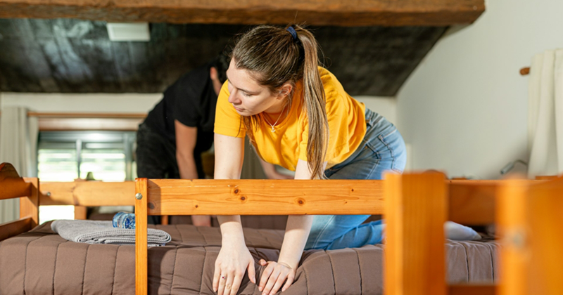 A woman leans over and adjusts the sheet on a bunk bed, with a man behind her