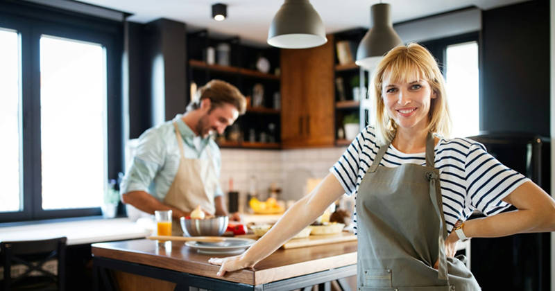 A woman stands next to a kitchen island with her hand on her hip while a man prepares food on it in the background