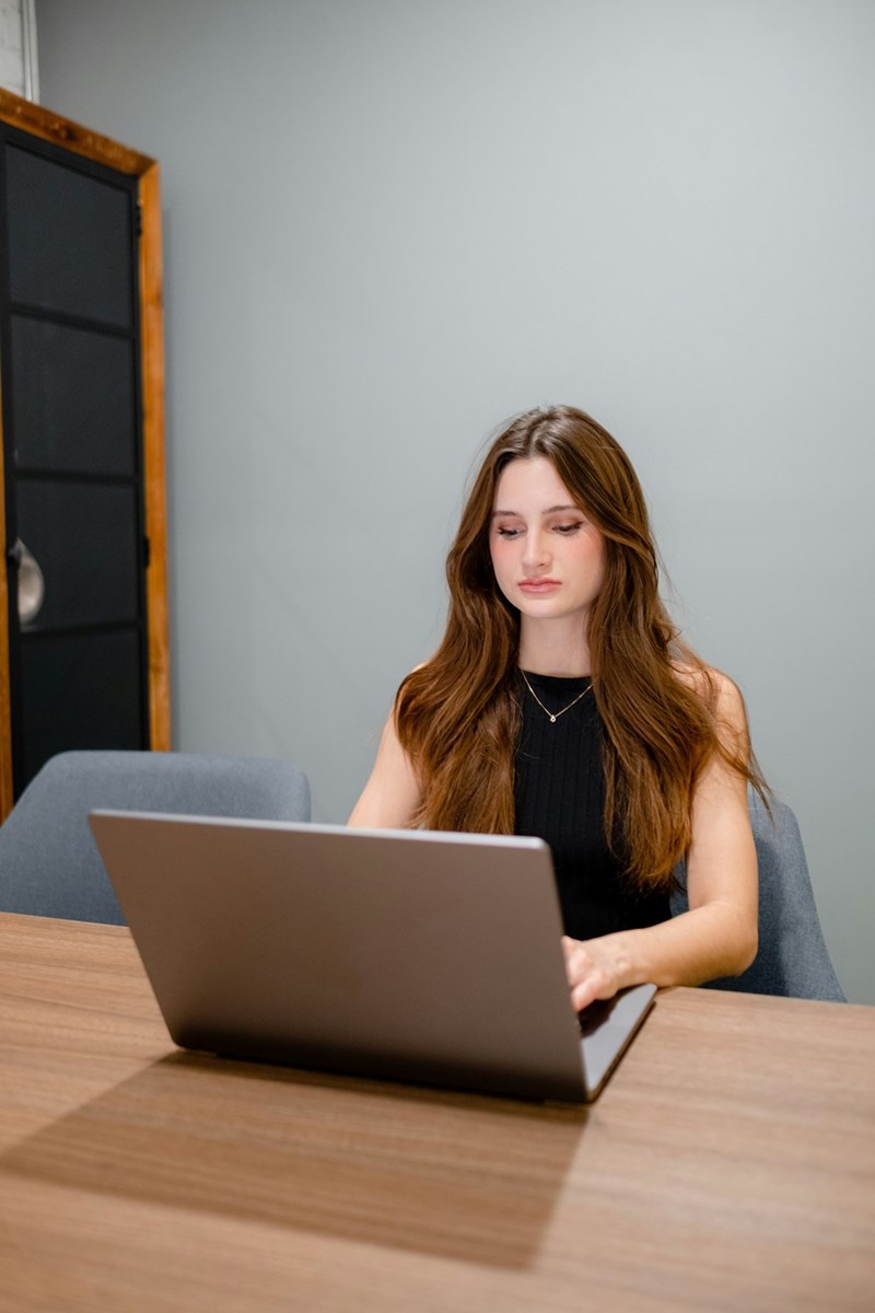 Woman sitting at a table working on a laptop in a minimal indoor setting.