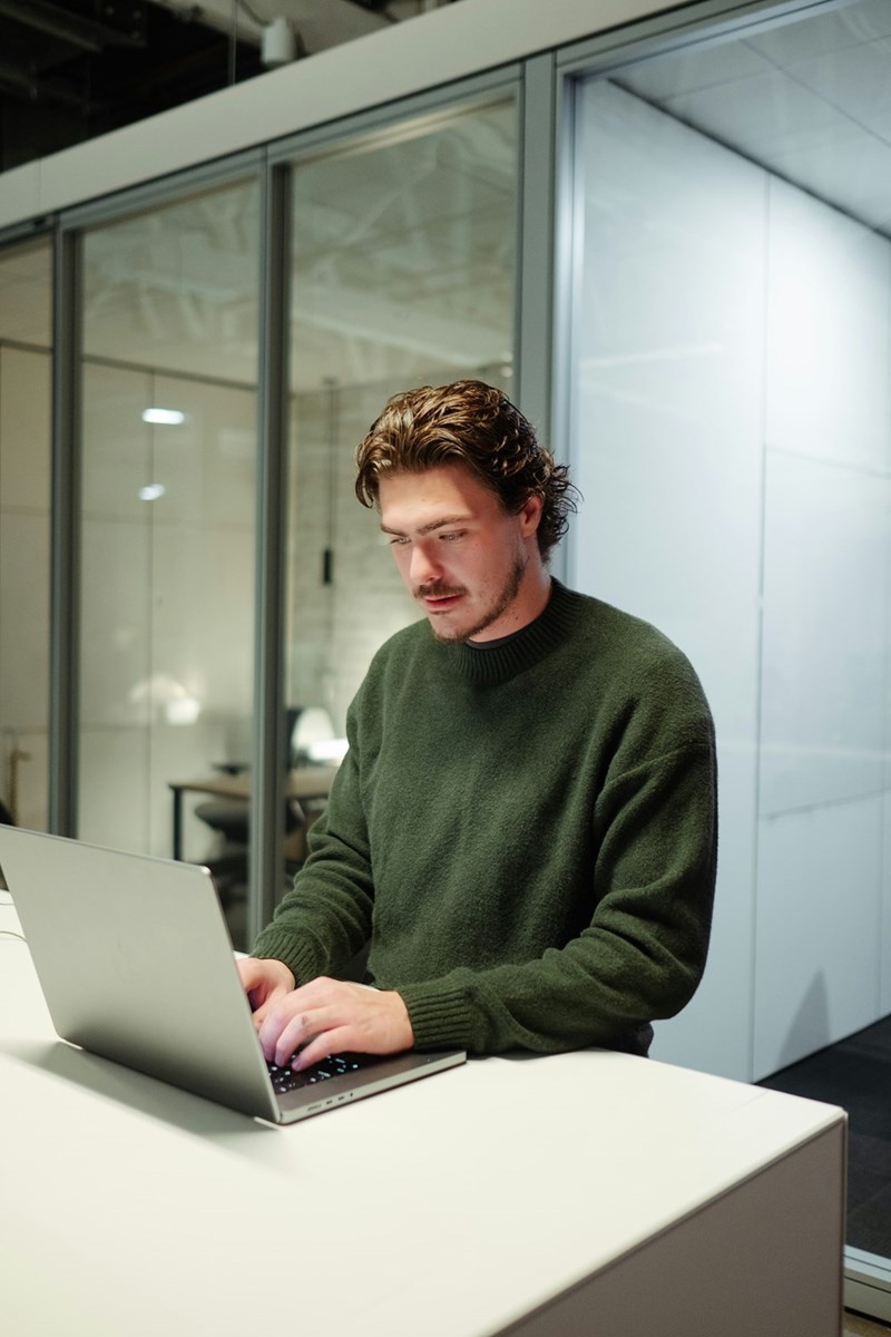 Man standing at a desk using a laptop in a modern office with glass walls.