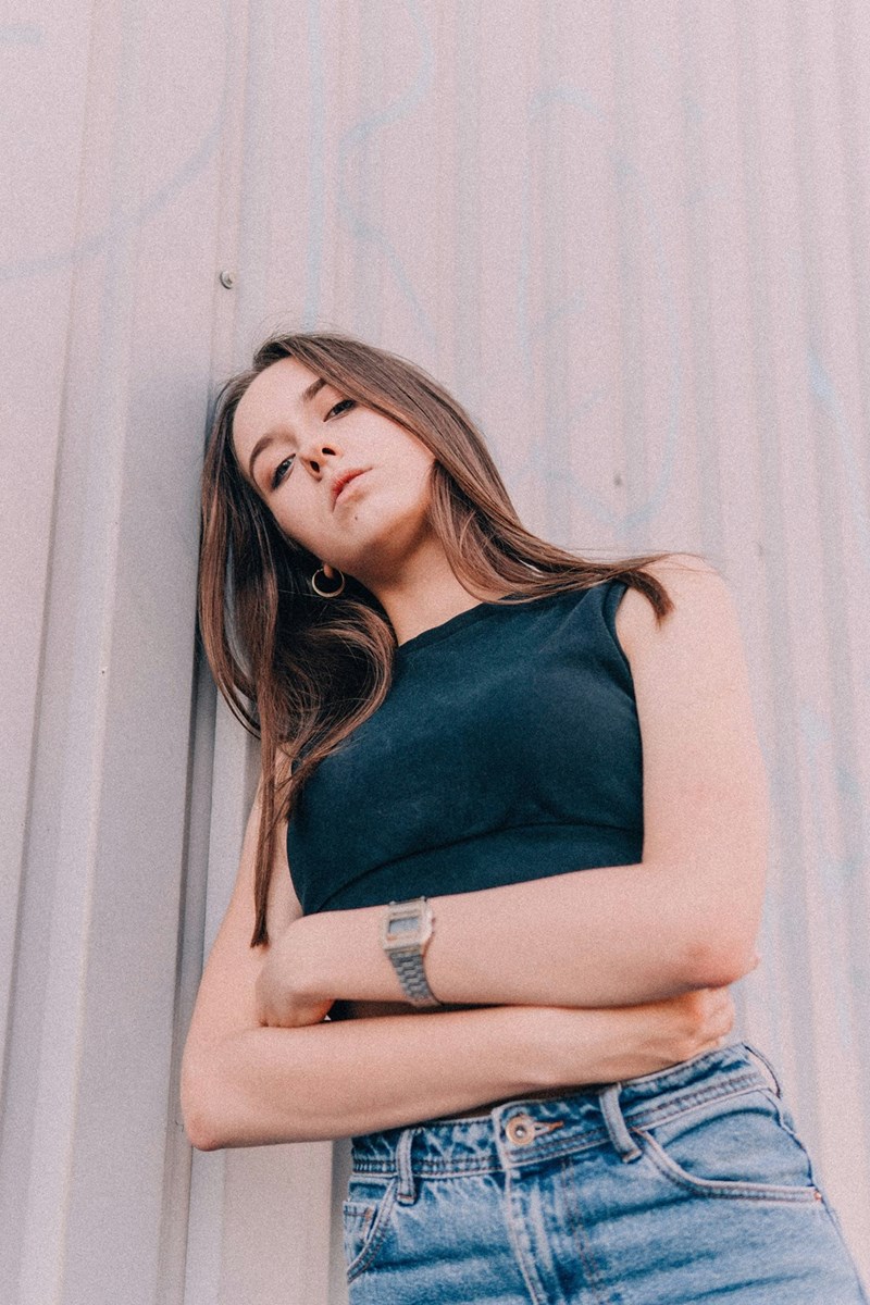 Young woman leaning against a wall with arms crossed, wearing a sleeveless top and jeans, looking at the camera.