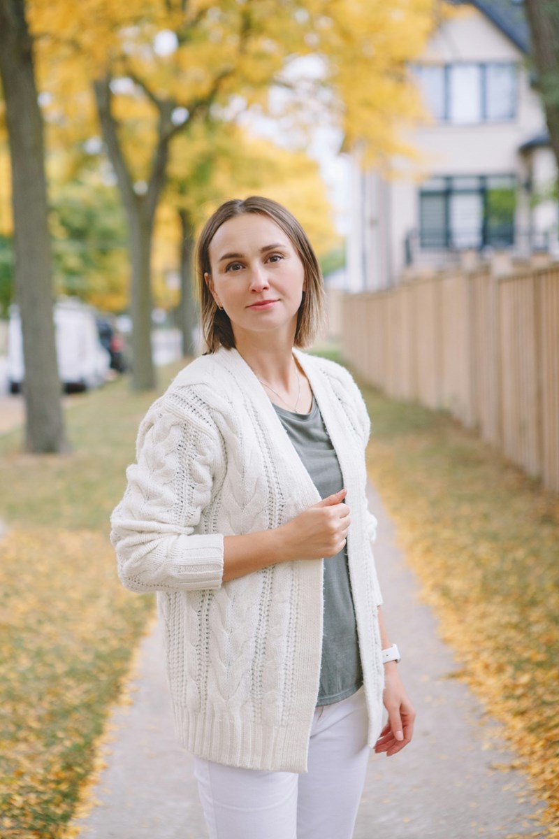 Woman standing on a tree-lined sidewalk in autumn, wearing a light cardigan and looking at the camera.