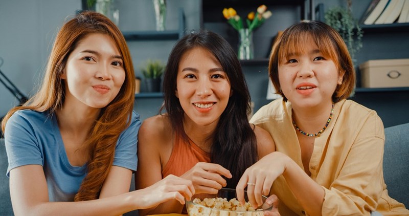 Three women smile and share popcorn while watching something entertaining.