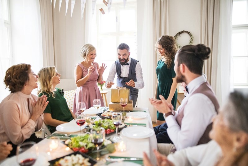 A group of people smile and clap as someone opens a gift during a joyful gathering.