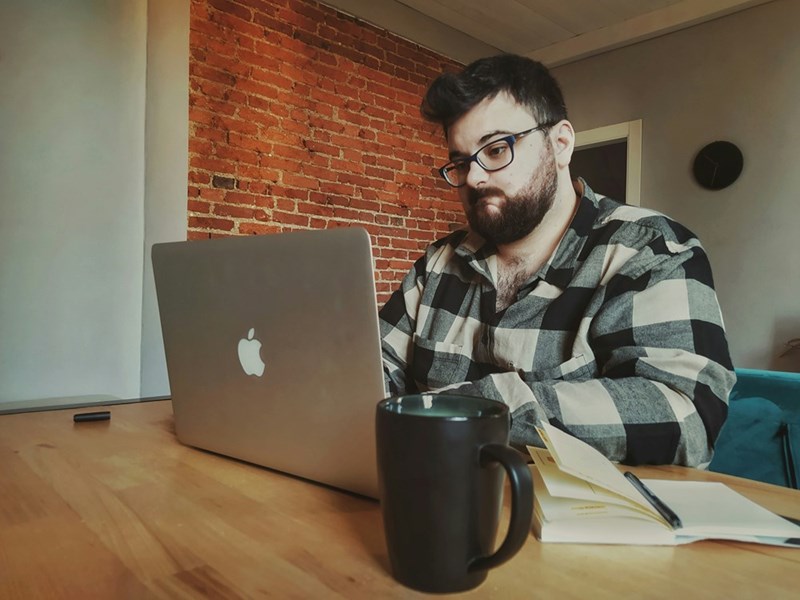 Man wearing glasses and a plaid shirt working on a laptop at a wooden table with a mug and notebook nearby.