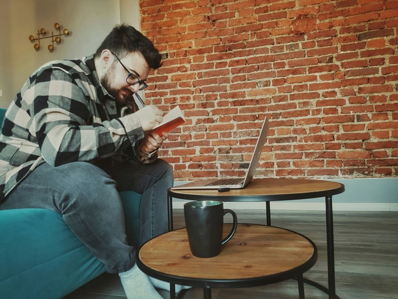 Man sitting on a couch writing in a notebook beside a laptop on a small table, with a mug in the foreground.