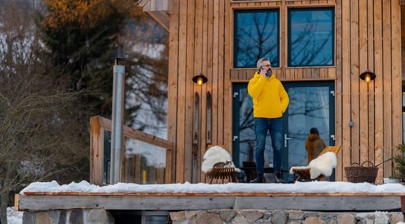 Man in a yellow jacket standing on a snowy deck outside a wooden house, talking on a phone.