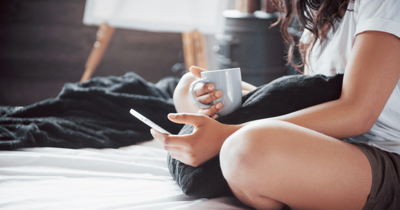 A woman sits cross legged in bed holding a mug and with a pillow between her knees, looking at her phone