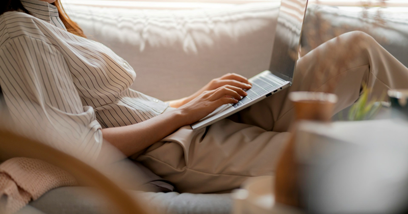 A woman wearing business casual reclines on a couch while using a laptop