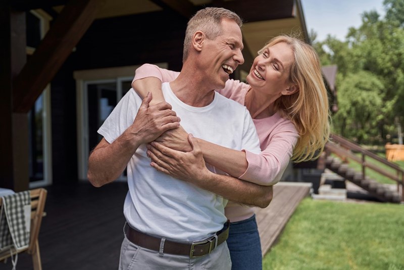 Waist-up portrait of happy woman hugging husband as they are standing in yard of countryside house