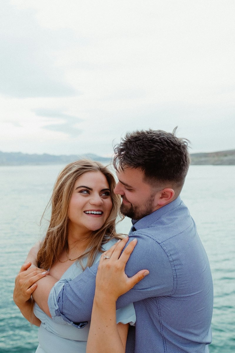 A man and a woman hugging by the water, showing off the woman's engagement ring