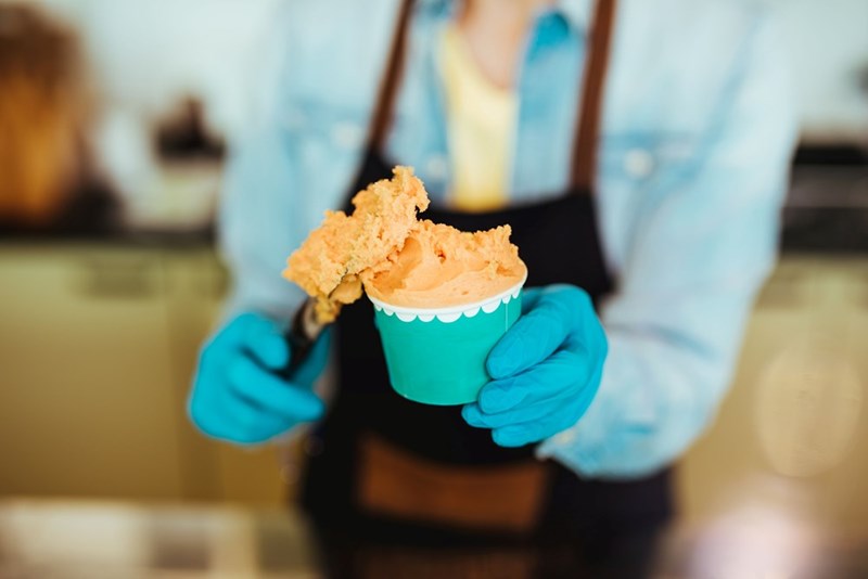 Gloved hands scooping orange ice cream into a small cup behind a counter.