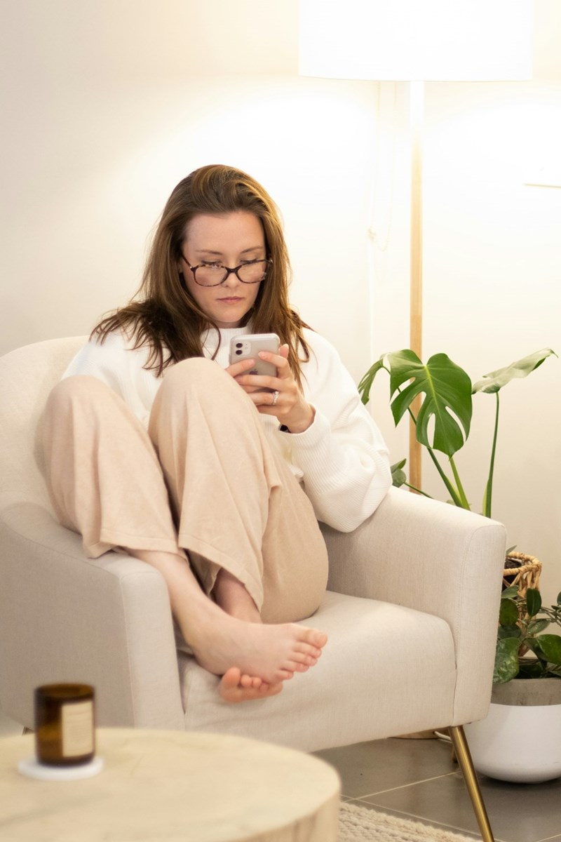 Woman sitting curled up in an armchair, wearing glasses and looking at her phone beside a floor lamp and plant.