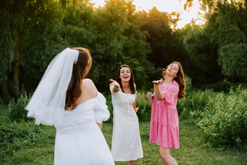 A bride shares a playful moment with friends as they blow kisses and laugh together in a sunlit green setting.